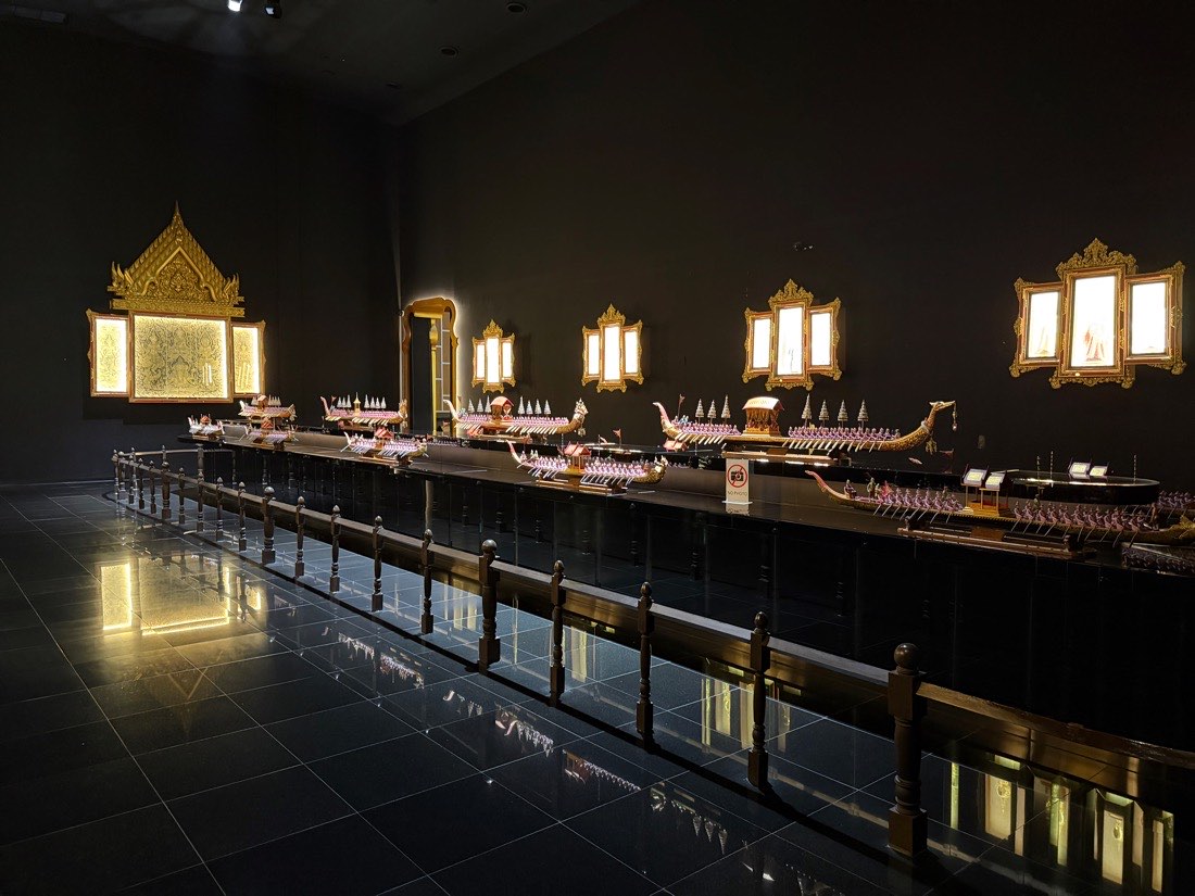 Models of Thai royal barges displayed in a darkened gallery under spotlights